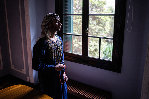 Young islamic man looking through window praying at home