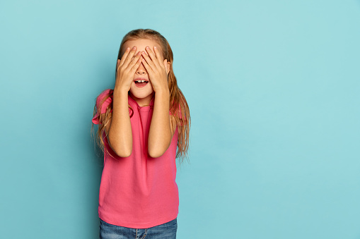 Portrait of little beautiful girl, child in pink T-shirt posing with hands covering eyes isolated on blue studio background. Hide and seek. Concept of childhood, emotions, facial expression, lifestyle