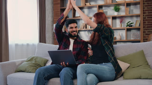 Happy overjoyed attractive young couple read good news and celebrating victory in online giveaway on laptop with raised hands and giving high five each other.