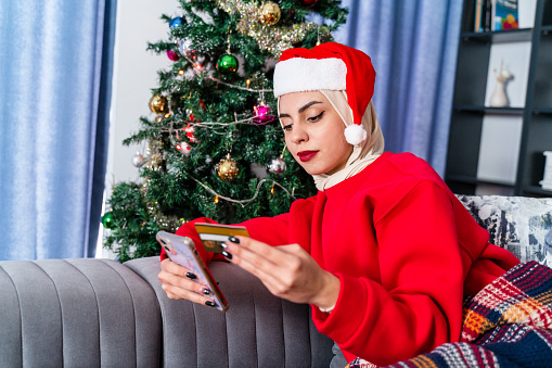 Happy female customer, shopper celebrating Christmas. modern Muslim Middle Eastern  girl in Santa hat using mobile phone for online shopping on winter December sale, paying for Xmas purchase, gifts by credit card