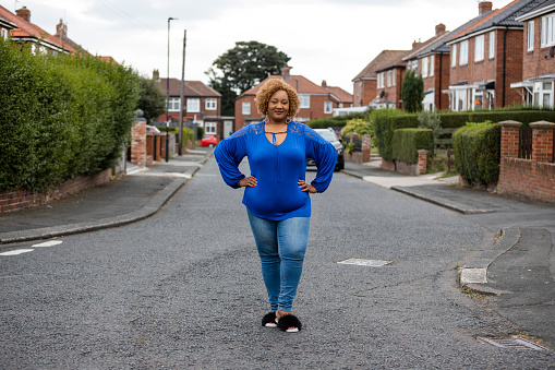 A portrait of a mid adult woman standing on a residential street in Newcastle Upon Tyne, England. She is looking at the camera and smiling with her hands on her hips.