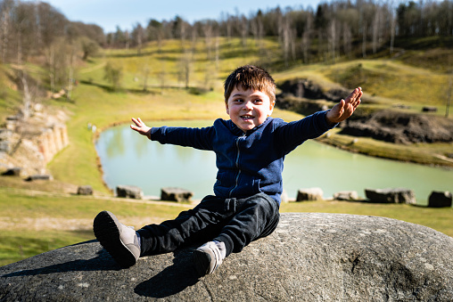Happy kid with open arms pretending to be a plane sitting on a boulder by a lake is engaging in a funny outdoor activity. Smiling child sitting like an airplane enjoying close contact with nature