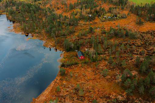 Drone photo of the mountain cabin by the lakeshore surrounded by the colorful autumn forest and the beautiful snowcapped mountain peaks in the Norwegian outdoors