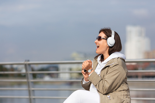 Attractive woman plus size sit on the city embankment and emotionally singing along to music with headphones, selective focus. Lifestyle, podcast, mental health, pleasure, privacy concept.
