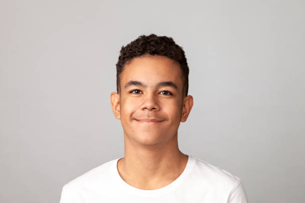 Close-up studio portrait of a cheerful 13 year old teenager boy in a white t-shirt against a gray background stock photo