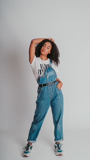 A portrait of a curly Hispanic female wearing denim overalls and posing against a wall