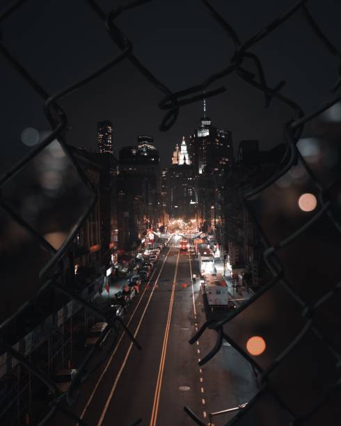 A view of a big long street in a huge city at night through a broken fence with skyscrapers in the background and many city lights