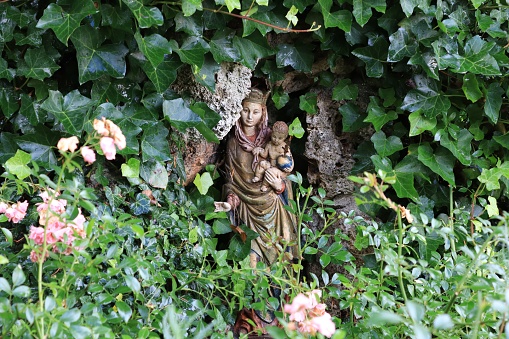A stone Statue of the Virgin Mary ringed by flowers and leaves in the garden