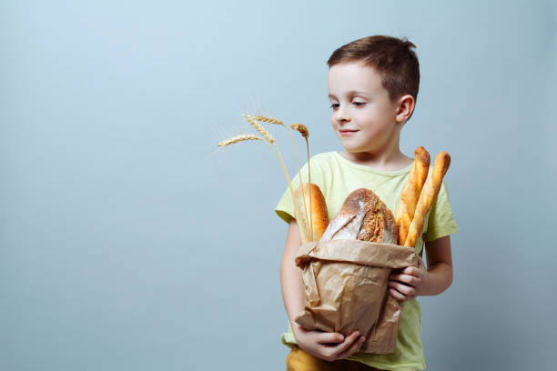 child with a grocery bag with freshly baked bread and ears of wheat, smiling softly against a uniform background. stock photo