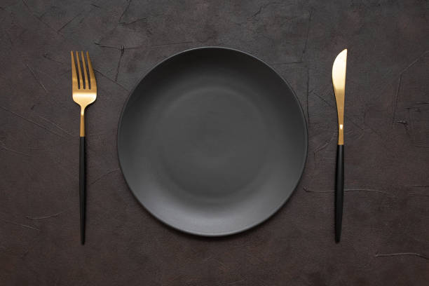 istock Closeup of a black plate with knife and fork on a black table 1440548872