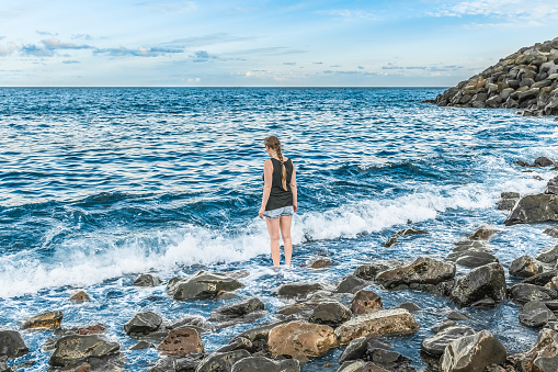 A lonely girl in clothes stands in the water and looks at the sea horizon - view from the back. A young woman among the waves and huge stones of a stormy ocean