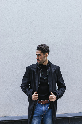 A vertical shot of a serious young man wearing a black coat, blouse, blue jeans with a necklace