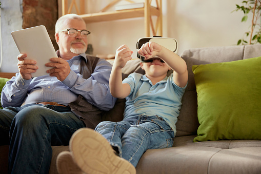 Modern gadgets. Portrait of happy senior man and little boy, grandfather and grandson using VR glasses sitting on sofa at home and laughing. Hobbies, leisure activities, family and emotions