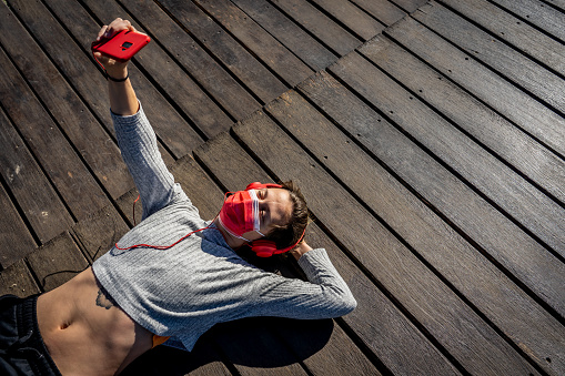 An overhead shot of an adult female with a facemask and headphones lying on a wooden dock and taking a selfie