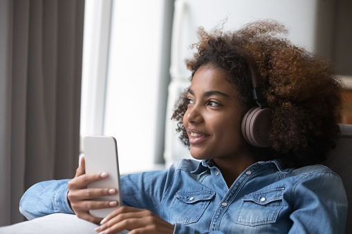 Close up African pretty girl wear headphones listens song spend time at home. Teenager using audio streaming and media services provider, enjoy favourite trendy track sit on sofa indoors. Hobby, tech