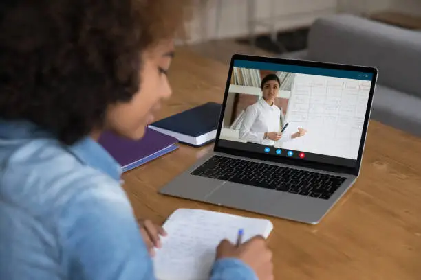 African teenager girl sit at desk with laptop, writes exercise, makes assignment, improve skills use modern tech, e-learning with online Indian teacher explain English languages tenses. Webinar event