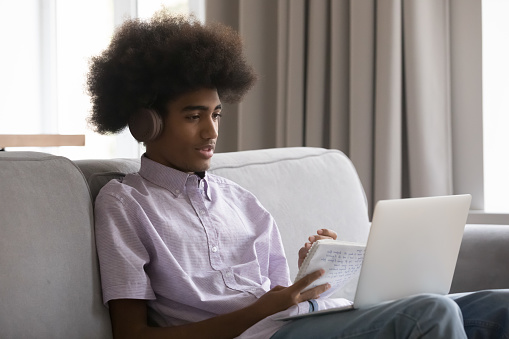 Attentive African student guy in headphones, holds copybook take notes, make assignment with classmate use video call, listen on-line tutor studying sit on sofa at home with laptop on laps. Education