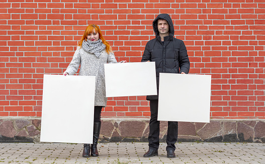 Young man and woman standing against a background of a red brick wall holding three white canvases in front of them to paint