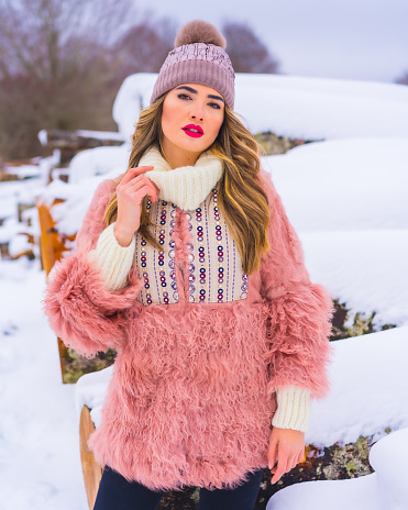 A vertical shot of a female wearing a pink fur jacket, pants, winter boots, and a hat in the snow