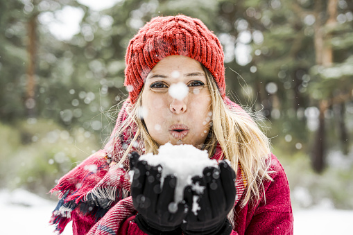 happy young woman playing with snow in winter, blowing snowflakes to camera, copy space