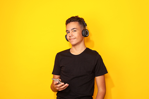 Studio portrait of a 13-year-old teenager boy in a black t-shirt with wireless headphones and a mobile phone against a yellow background