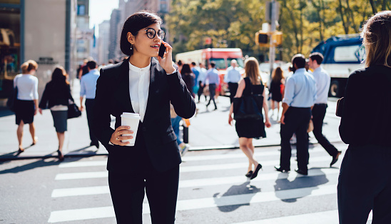 Modern female in formal clothes and eyeglasses standing on street and talking on smartphone while drinking hot takeaway coffee