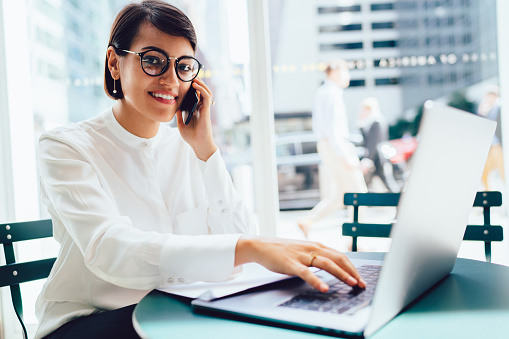 Smiling elegant female in formal clothes and eyeglasses sitting in modern office while working on laptop and talking on smartphone