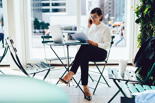 Full body cheerful female in formal clothes and eyeglasses sitting in modern office while working with papers and talking on smartphone