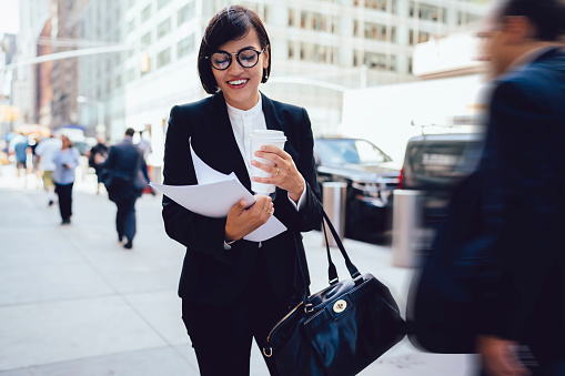 Content young businesswoman with black hair in formal work attire in eyeglasses walking on pavement holding takeaway cup and documents
