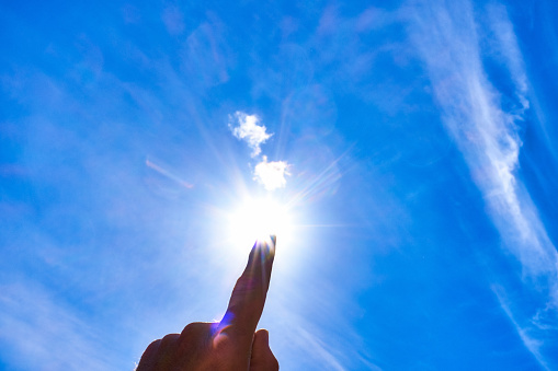 Man pointing upward at the bright sun in a blue sky streaked with cloud.
