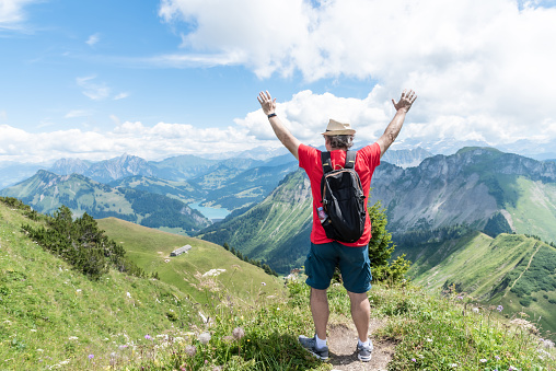 Senior adult man with hat, backpack and red shirt with his arms up looking towards the Swiss Alps in the background