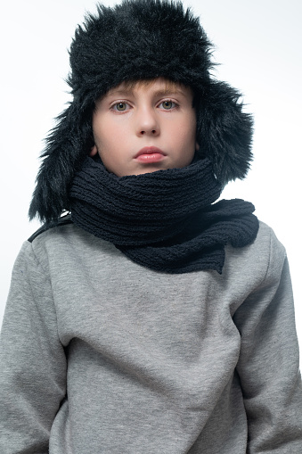 Portrait of a child in a winter hat with earflaps and a knitted scarf on a white background, black winter clothes.