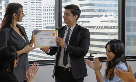 Young caucasian manager in suit giving mock up certificate to woman staff with happy smiling face while others applauding for congratulation in office meeting room. Selective focus.