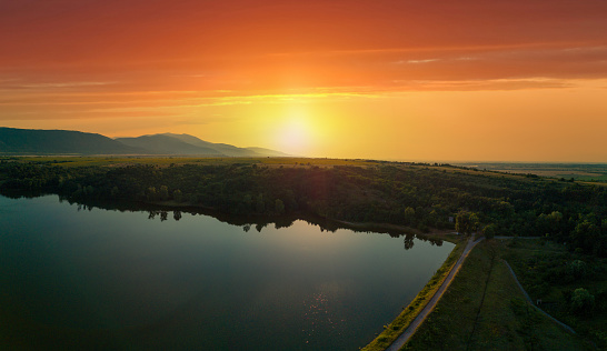 Bright sunset yellow-orange sun illuminates agricultural fields, high mountain silhouettes of Balkan mountains and small lake. Panorama, top view
