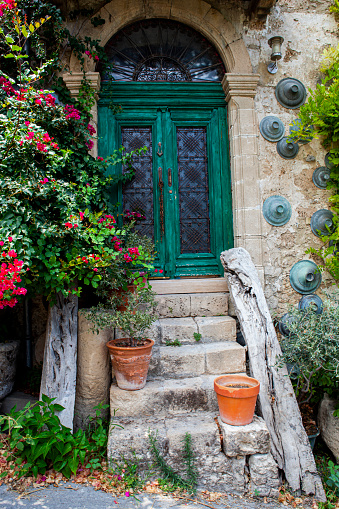 Natural stone wall, wooden door, green shuttered window with glass panes. Potted plants with red flowers. On a cobblestone street.