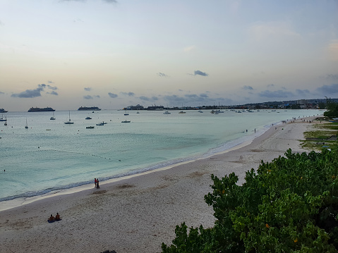 beautiful tropical beach with blue water in the ocean, Barbados, unrecognized people walking on sand from distance. View on port, ships, vessels, cruises and fishing boats in harbor