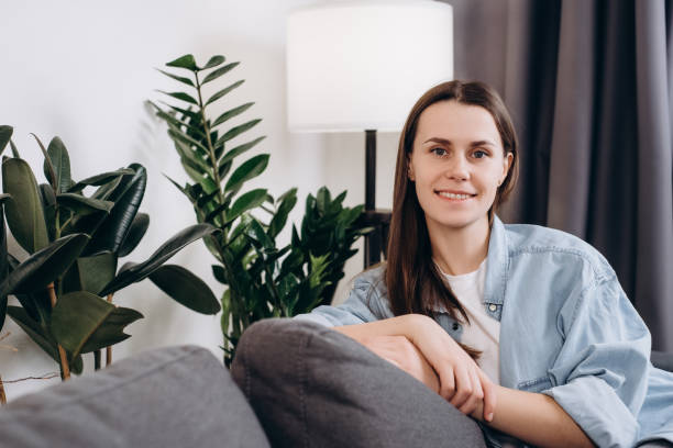 mulher jovem feliz sentada no sofá em casa, sorrindo olhando para a câmera. retrato da menina adorável 20s anos de idade relaxando no sofá. alegre bela morena feminina e relaxante durante o outono ou inverno - 20s 20 25 years beauty beautiful - fotografias e filmes do acervo