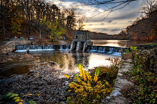 Morristown, NJ - USA - Nov 5, 2022 An autumnal horizontal wide angle view of New Jersey's historic stone Speedwell Dam during sunset. Fly fisherman in the foreground and people hiking in the distance.