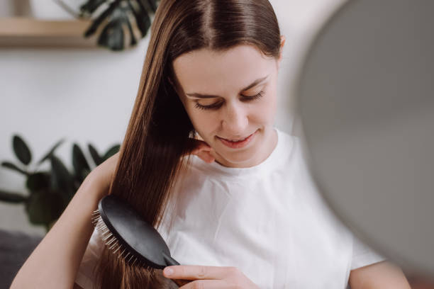 atraente sorridente jovem morena anos de idade 20 anos sentada no sofá confortável na sala de estar em casa, penteando seu belo cabelo sedoso com escova de madeira. cuidados com os cabelos, pessoas e conceito de estilo de vida - 20s 20 25 years beauty beautiful - fotografias e filmes do acervo