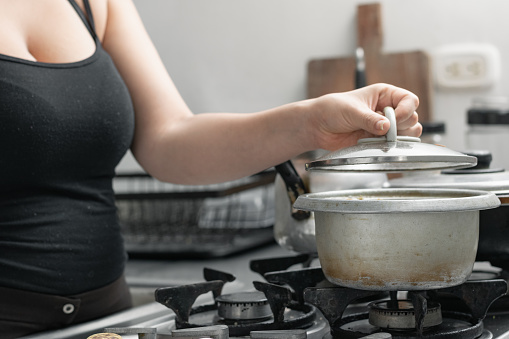 young woman uncovering a pot on the gas stove, lifting the lid to see the food she is preparing.