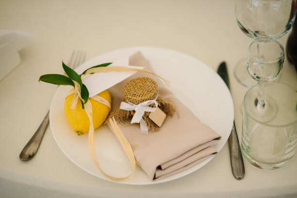 istock Serving, setting table. Gift citrus lemons decorated with ribbons and branch greenery on plate. Compliment for guests. Banquet table served with silverware, plates with napkins, cutlery and glasses. 1440592346