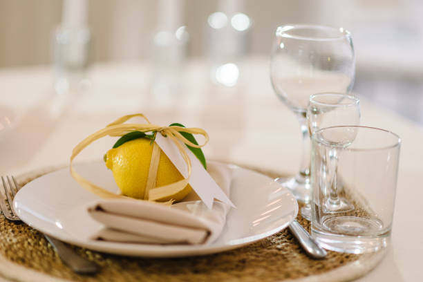 istock Serving, setting table. Gift citrus lemons decorated with ribbons and branch greenery on plate. Compliment for guests. Banquet table served with silverware, plates with napkins, cutlery and glasses. 1440592362