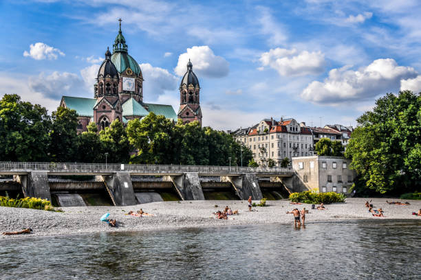 People Enjoying Sunny Day On Isar River Beach Beneath St. Lukas Cathedral In Munich, Germany