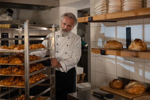 A gray-haired bearded baker in a white baker's uniform prepares dough, mixes flour, eggs  in the morning rays filtering through the window in the bakery. The baker smells intoxicating aromas of freshly baked French croissants.