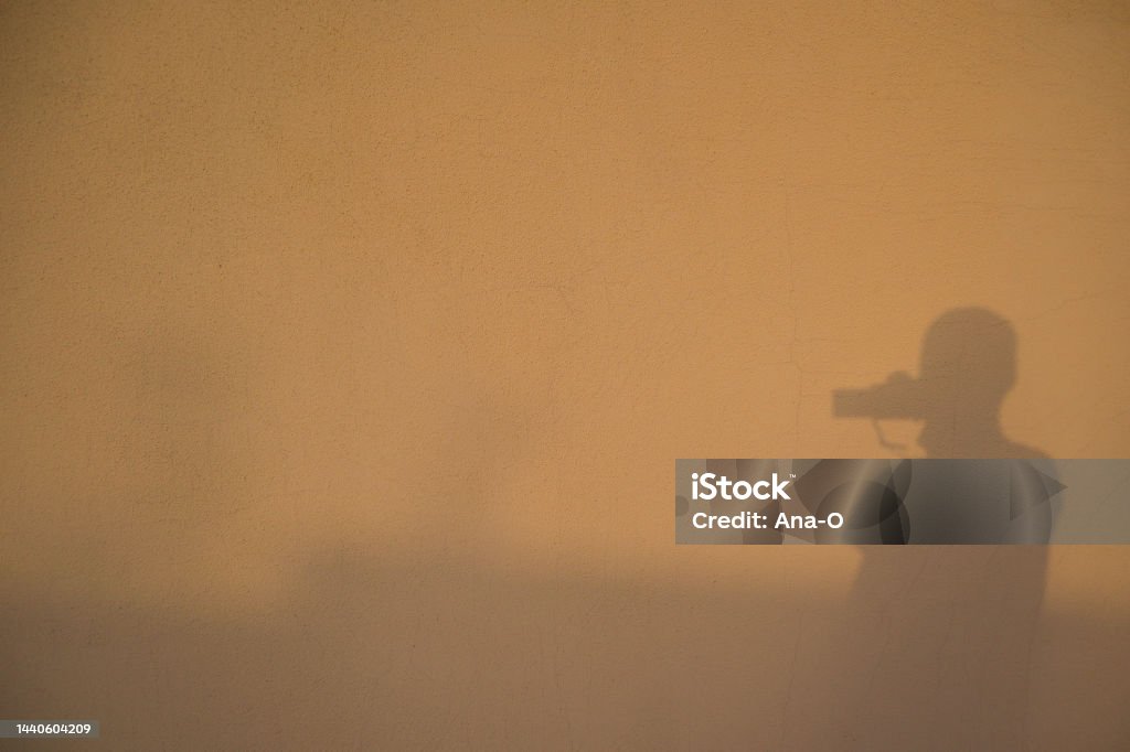Shadows in a wall. Reflection / silhouette of the photographer taking photo of himself. Silhouette of photographer taking a photo Shadows in a wall. Reflection / silhouette of the photographer taking photo of himself. Black shadow silhouette of young man with photo camera Art Stock Photo