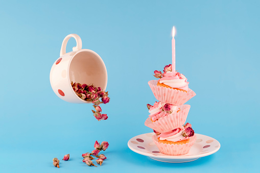 cup with flowers of rose and  three birthday  berry muffins  on top of each other in pink wrappers with a candle on top on a saucer on  blue background  high-quality photos for calendar and cards.