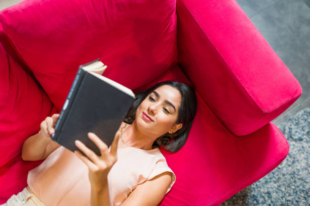 Top view of a relaxed young woman resting on the sofa while reading an interesting book
