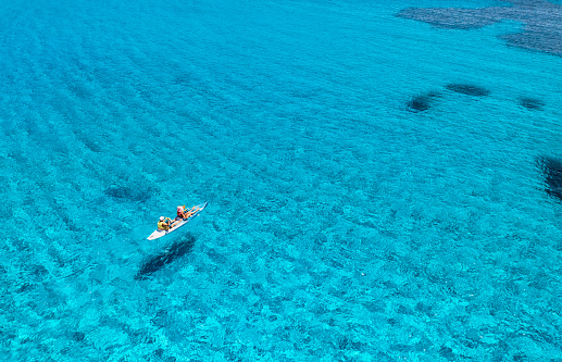 Aerial view of kayak with people in blue sea at sunset in summer. Man and woman on floating canoe in clear azure water. Sardinia island, Italy. Tropical landscape. Sup board. Active travel. Top view