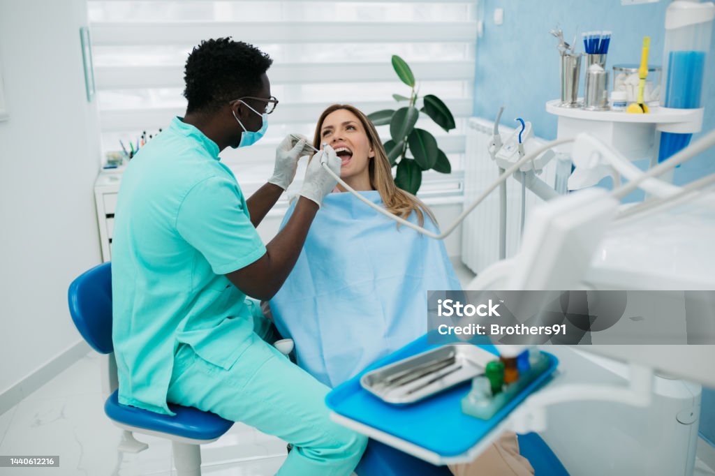 A professional dentist carefully working with his female patient Young adult female patient comfortably sitting in a dentist chair with her mouth open while her dentist is sitting next to her and using his work tools to carefully inspect her teeth Dentist Stock Photo