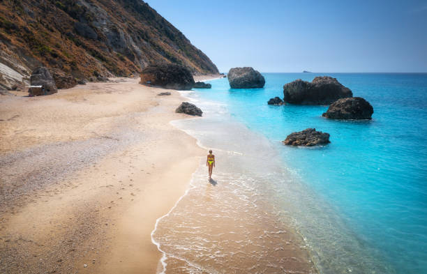 해질녘에 파도가 있는 바다 근처의 모래 해변에서 걷는 젊은 여성의 공중 전망. 그리스 레프카다 섬에서의 여름 휴가. 스포티 한 소녀, 푸른 물, 산의 최고 전망. 라이프 스타일과 여행 - beach blue turquoise sea 뉴스 사진 이미지